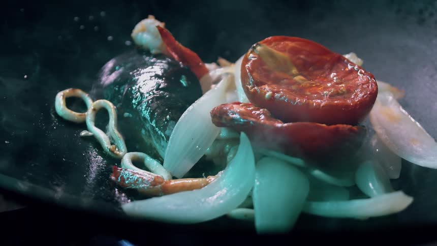 The video displays a closeup of various ingredients including mussels shrimp sliced onions and halved cherry tomatoes cooking in a dark pan the steam rising from the pan indicates the ingredients are actively heating