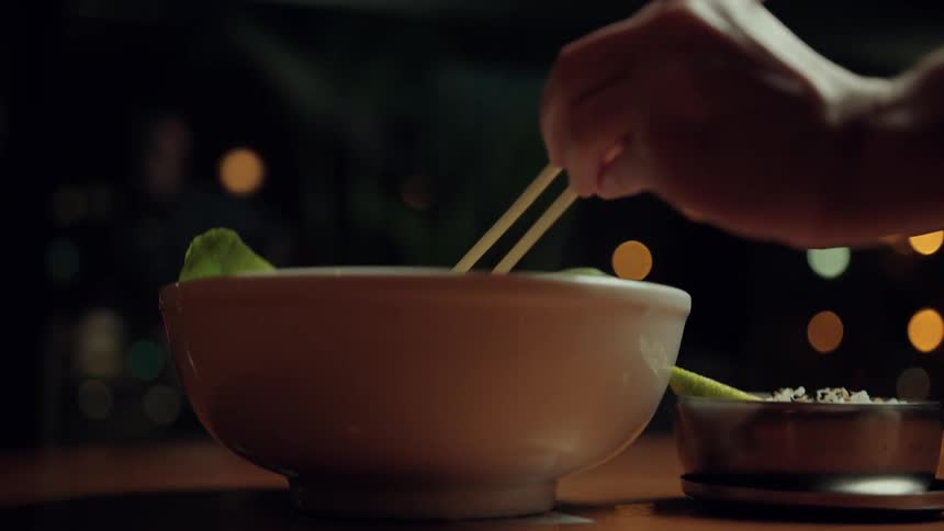A hand holding chopsticks lifts a bite of food from a bowl with soft bokeh lights glowing in the background