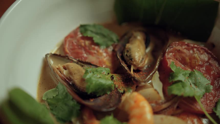 A close view of a bowl of hot soup shows large green leaves mussels shrimp and red tomato slices in a light brown broth garnished with fresh green cilantro
