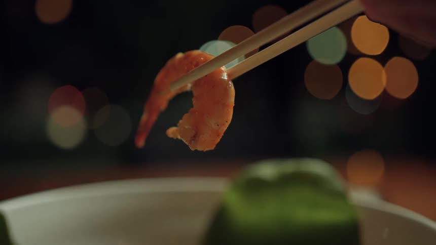 Shrimp held by chopsticks above a bowl in low light
