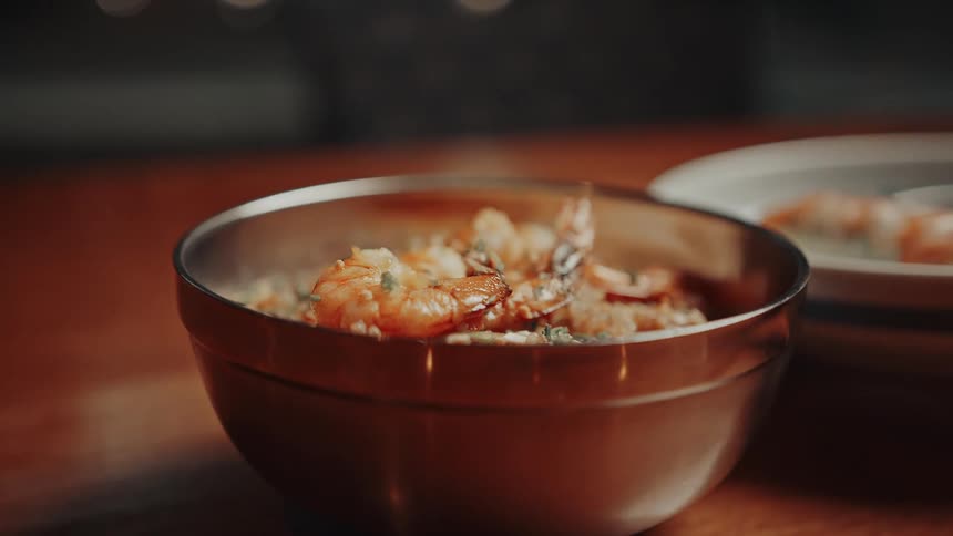 A close view shows a steaming metal bowl filled with fried rice and shrimp resting on a wooden table with another similar bowl blurred in the background