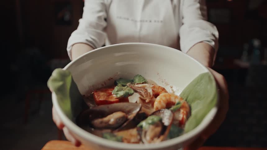 A chef presents a bowl of seafood stew in a warmly lit restaurant