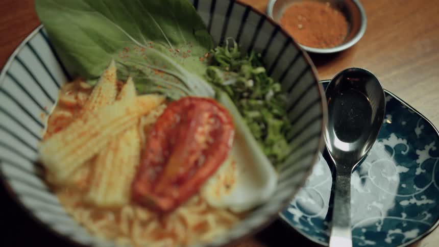A bowl of ramen noodles with baby corn tomato greens and beside a spoon and spice dish