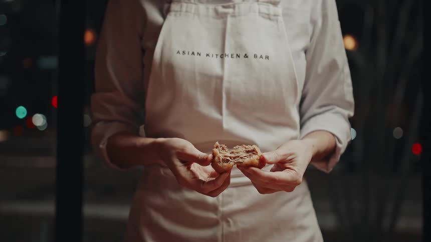 A chef in an asian kitchen bar apron holds a freshly broken piece of food possibly highlighting texture or filling