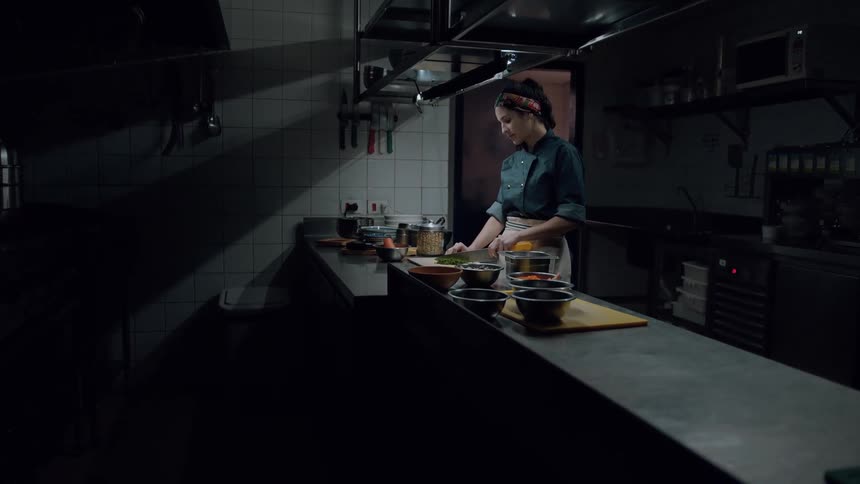 A female chef chops vegetables at a counter in a dimly lit kitchen