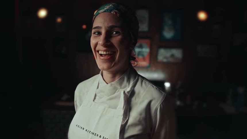 A smiling chef stands indoors wearing a white apron and headscarf