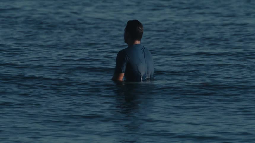 A man stands in the ocean near the shore at midday with half of his body submerged
