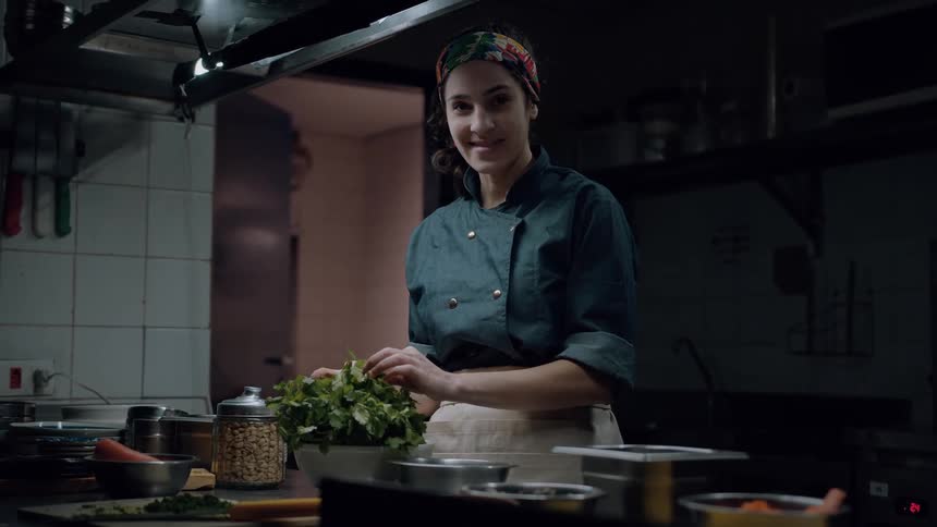 A young chef wearing a dark green chefs coat and a colorful headscarf smiles at the viewer while preparing green herbs on a metal counter in a dimly lit kitchen