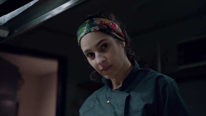 A female chef looks into the camera in a dimly lit kitchen