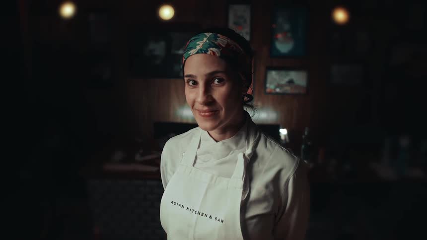 A smiling female chef stands in a dimly lit restaurant kitchen