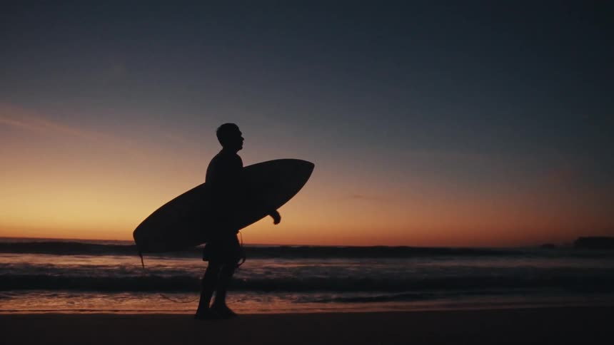 A silhouette of a surfer walks along the beach at sunset board in hand