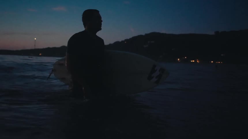 A male surfer walks out the ocean with his surfboard at his side looking towards a dark shoreline with lights in the distance under a twilight sky