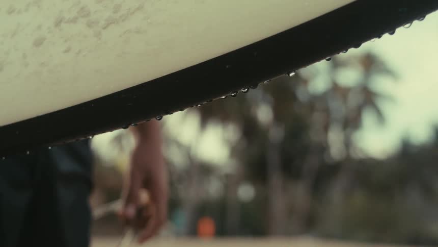 A man holds a surfboard while standing on the beach at midday with water dripping from the board