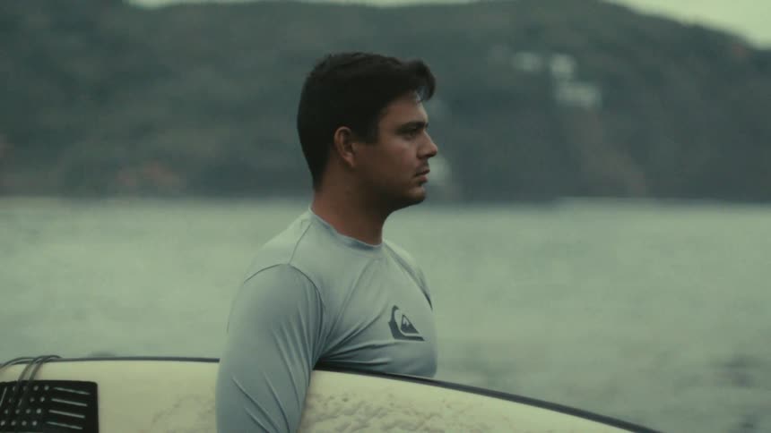 A surfer stands by the water holding his board gazing toward the ocean with a calm expression