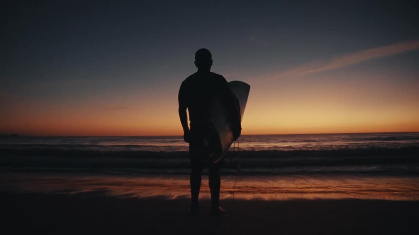 A lone surfer stands silhouetted against a vibrant sunset on a beach holding a surfboard and gazing out at the calm ocean waves