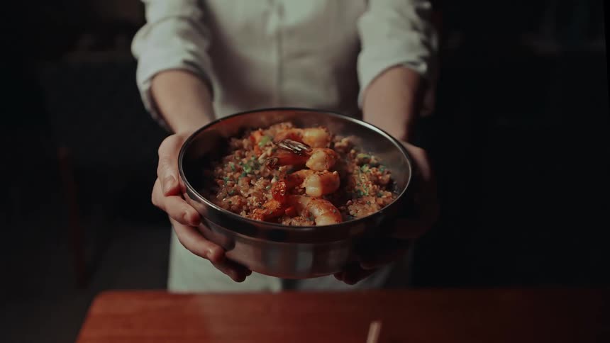 A chef in a white apron presents a bowl of colorful food holding it out with both hands in a warmly lit setting