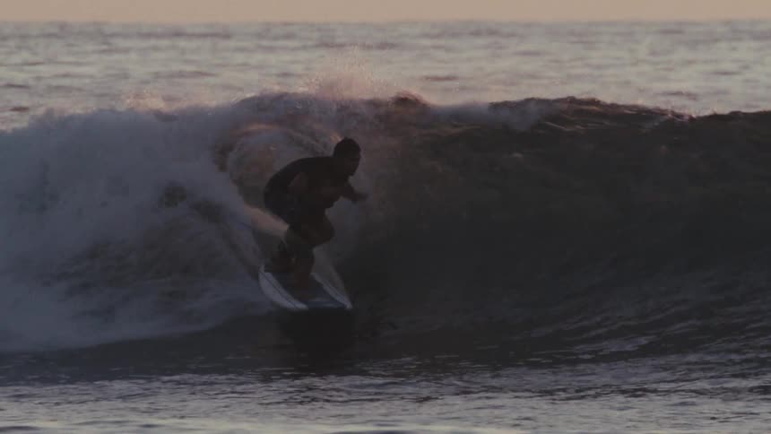 A man and a child ride a wave together on a surfboard their silhouettes defined against a dusky sky as foaming water churns around them