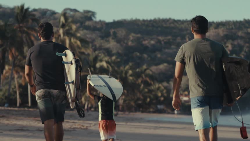 Two men and a boy walk along the beach while carrying surfboards during daytime