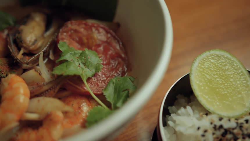 A bowl of steamed white rice is topped with black and white sesame seeds and garnished with a slice of lime is next to a seafood stew