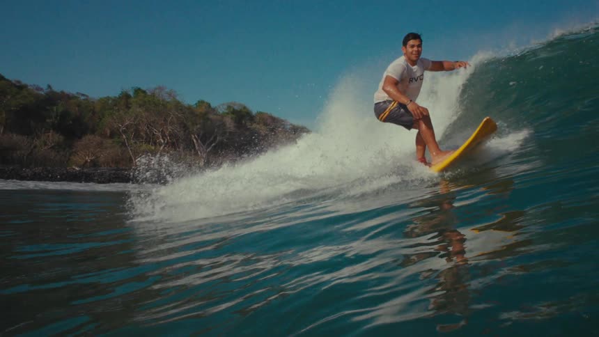 A young adult male surfer on a yellow surfboard rides a large ocean wave with lush green trees lining the distant shore under a clear blue sky