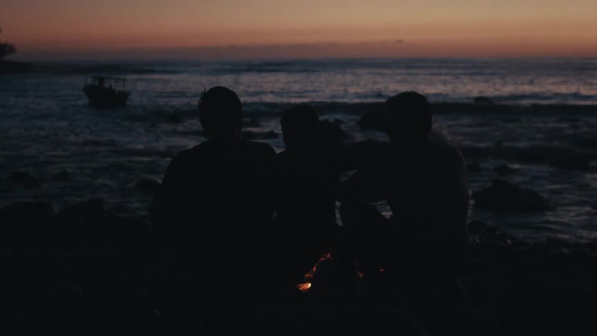Silhouette of friends sitting near a campfire next to the shoreline watching the sunset