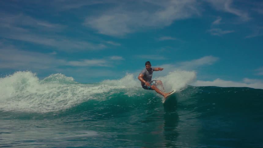 Male surfer rides a breaking wave on a white surfboard under a cloudy summer sky