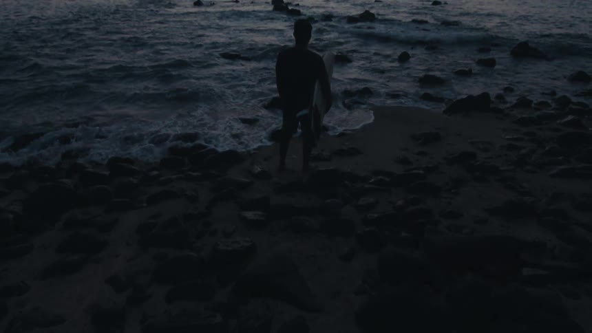 A man stands on a rocky beach at sunset watching the waves crash against the rocks