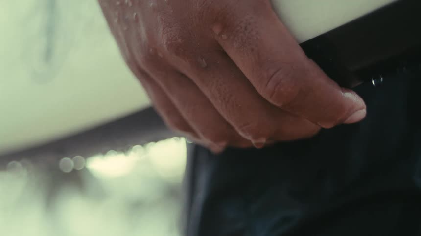 A close view shows a persons hand holding the edge of a surfboard with water droplets dripping from their fingers against a blurred light background