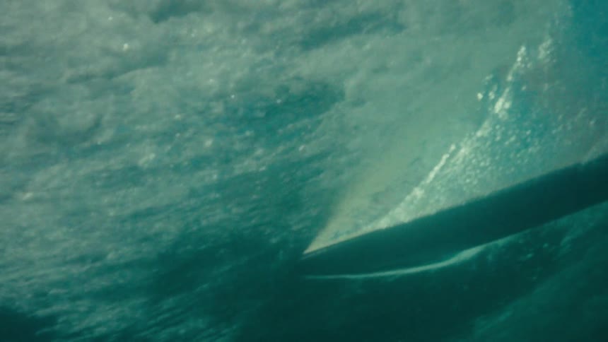 A young male surfer on a black surfboard rides a large ocean wave then the camera submerges underwater with bubbles and the underside of the board visible in the turquoise water