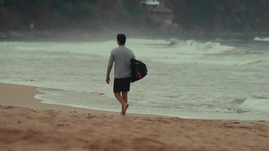 The man is walking on the beach while carrying a bag