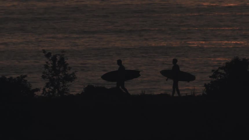 The video displays the silhouettes of two surfers standing on a beach or cliff at sunset holding their surfboards the sun has already dipped below the horizon casting an orange and purple glow over the calm ocean waters