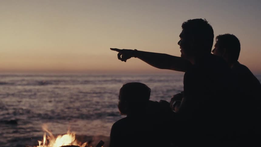 The video shows the silhouettes of three people gathered around a campfire on a beach at dusk one person in the foreground points out towards the calm ocean illuminated by the fading light