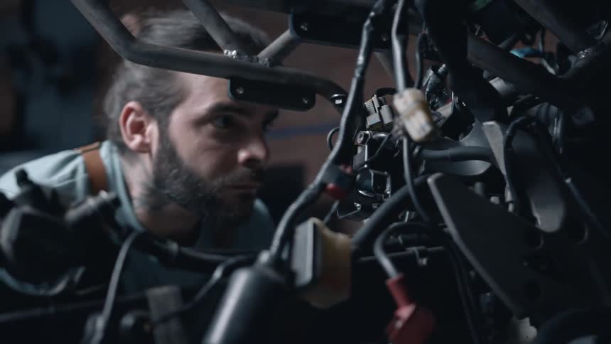 A focused mechanic with a beard and ponytail leaning in to meticulously work on the complex wiring and components of a motorcycle