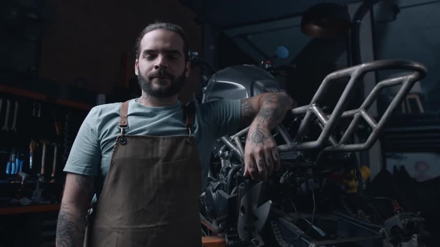 A tattooed mechanic in a brown apron stands confidently beside a partially assembled motorcycle in a dimly lit workshop