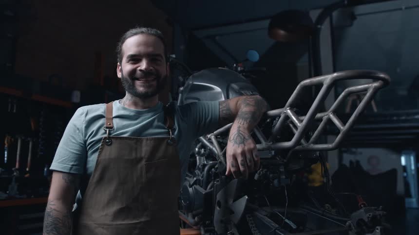 Man smiles next to a disassembled motorcycle in a mechanical workshop