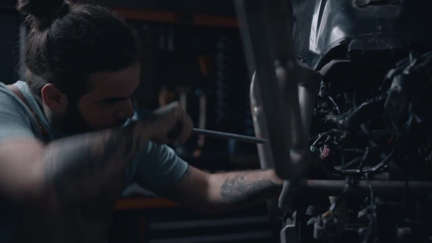 A tattooed mechanic uses a screwdriver to work on the wiring of a motorcycle in a dimly lit workshop