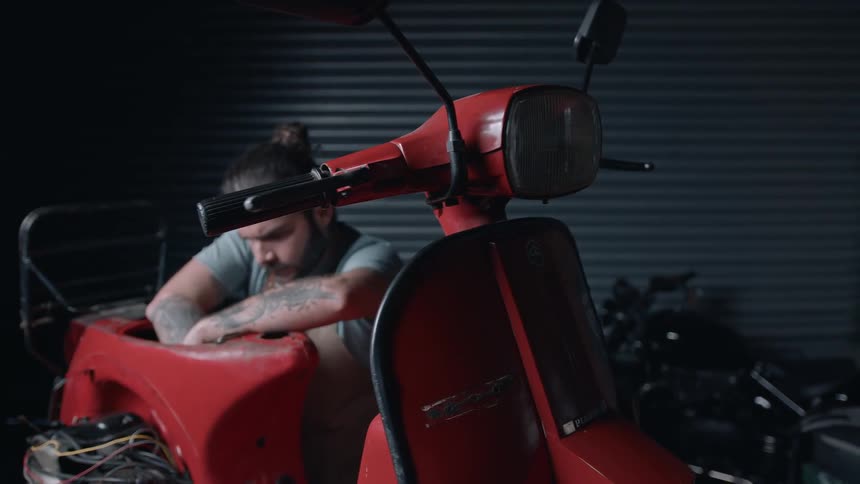 A bearded man reaches into the interior of a bright red motor scooter in a garage