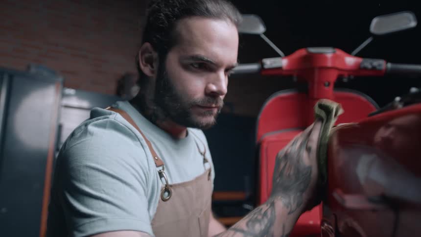 A focused mechanic polishes a red scooter with a cloth in a welllit garage