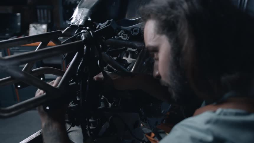An adult man with a beard and long hair works intently on the engine of a dark motorcycle surrounded by tools and equipment in a garage