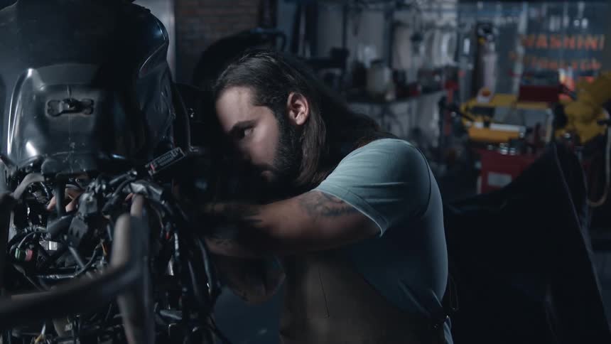 An adult man with long hair and a beard wearing a light colored shirt leans over the engine of a motorcycle working intently on its internal components in a dimly lit garage