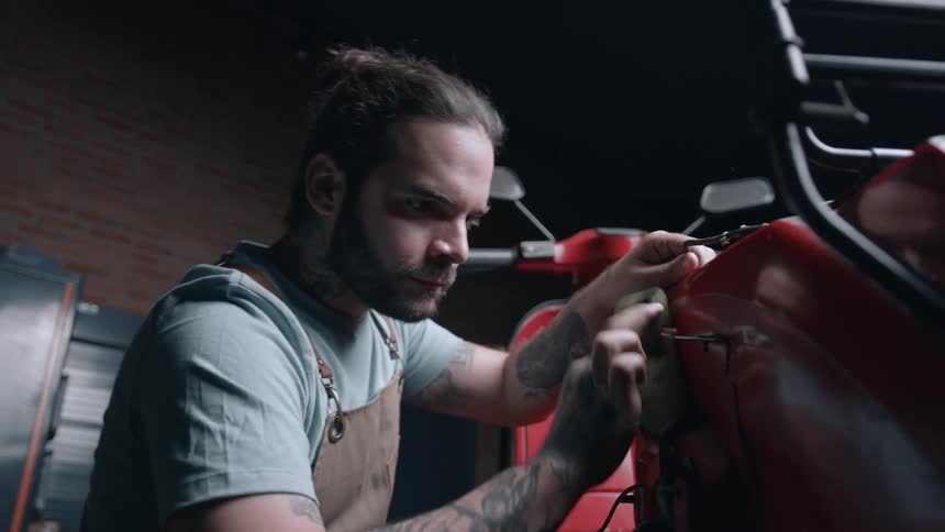 Close up of a young male mechanic wiping the body of a red motorcycle with a cloth in a workshop