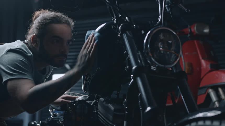 A young adult man with a beard and hair tied back wearing a light colored shirt leans over a dark motorcycle carefully cleaning its fuel tank with a cloth in a garage setting