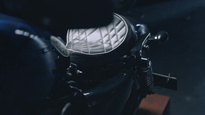 A sleek motorcycle features a quilted white leather seat highlighted under focused lighting in a dark garage