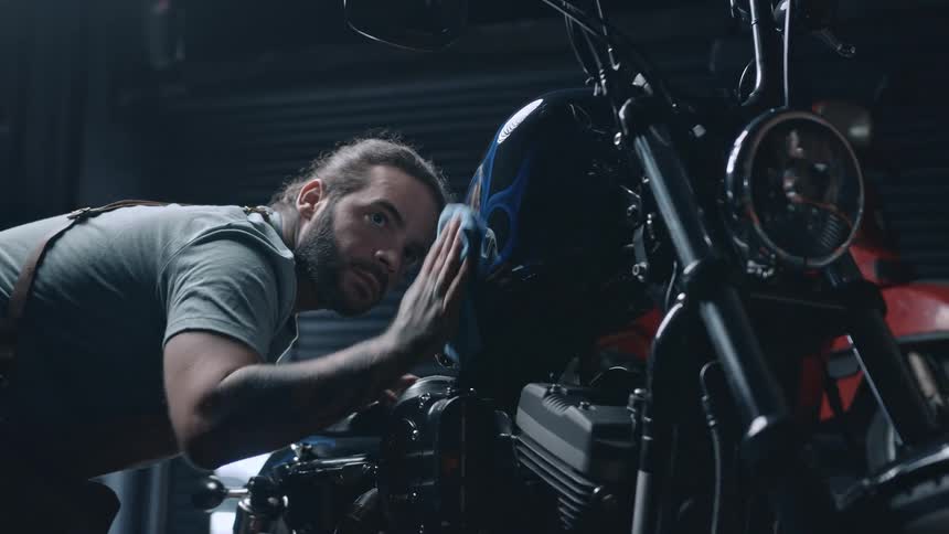 A young male mechanic wipes a motorcycle body with a cloth in a workshop during daytime