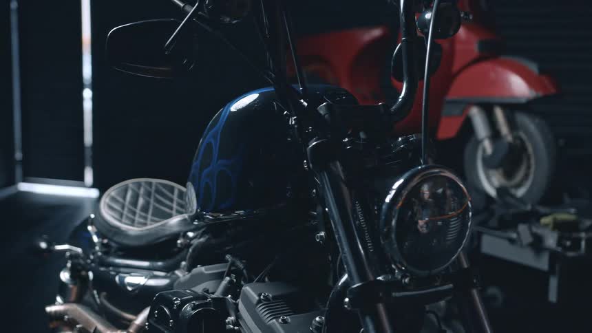 A shiny black motorcycle with blue flames painted on its fuel tank is parked in a garage with a red scooter visible in the background