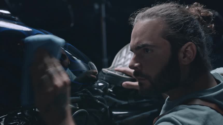 A man with a beard and hair tied back is meticulously polishing the dark reflective surface of a motorcycle with a blue cloth in what appears to be a dimly lit setting