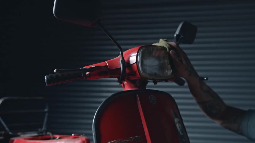 A man thoroughly wipes the headlight of a red motor scooter