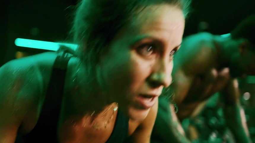 A focused closeup of a determined sweating woman engaging in an intense indoor cycling workout under atmospheric green lighting and a man also doing workout in the back