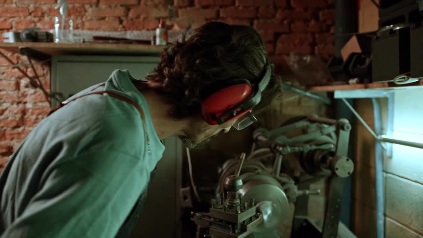 A man inside a workshop wears headphones and safety glasses while working with tools