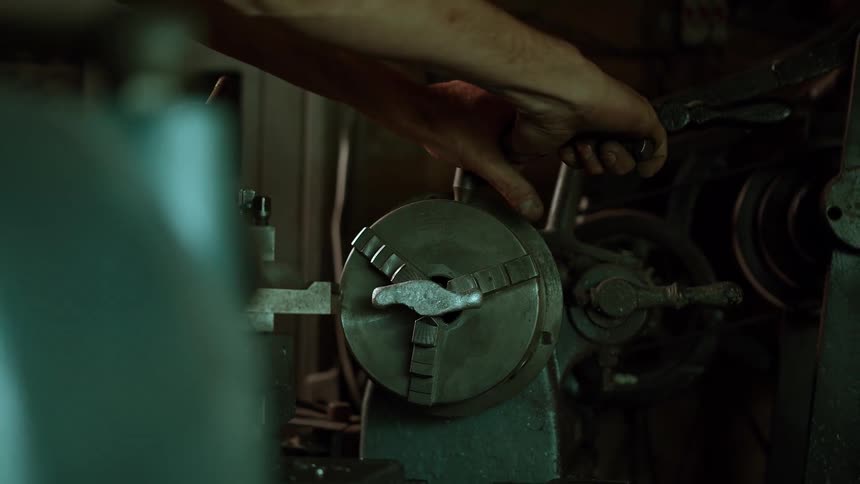 Hands adjust the chuck of a lathe machine in a dimly lit workshop